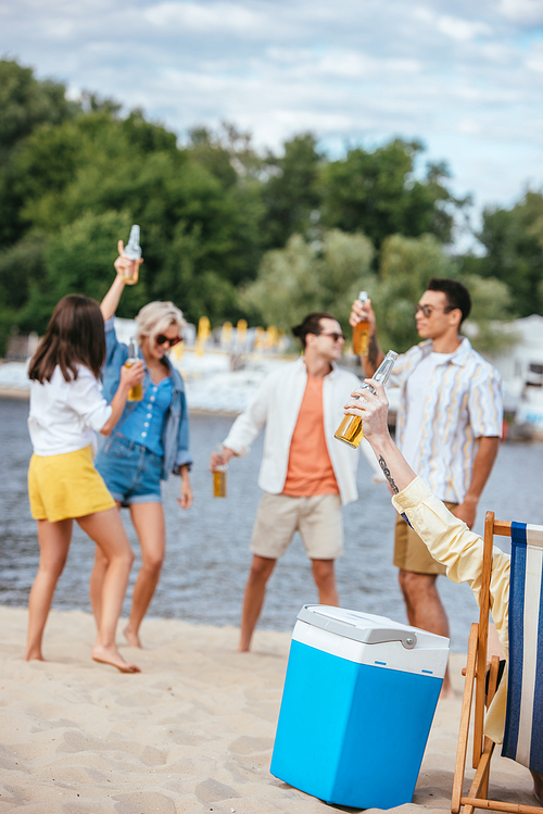 selective focus of cheerful multicultural friends holding bottles of beer while having fun on beach