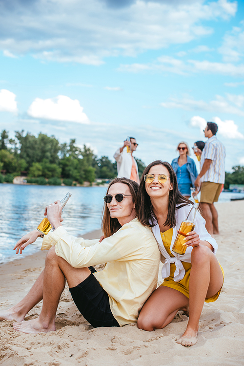 cheerful young couple sitting on sand and holding bottles of beer near multicultural friends