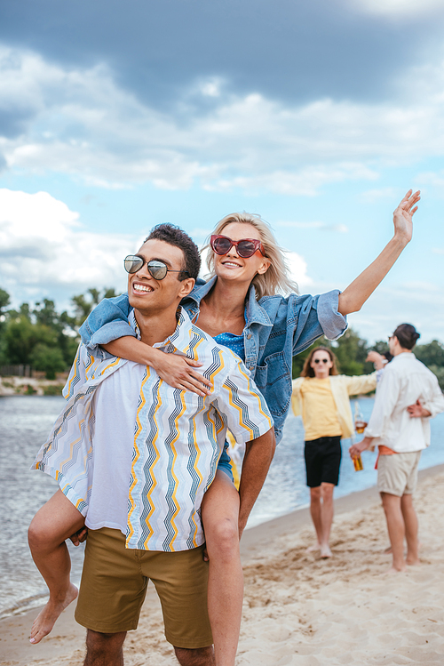 smiling mixed race man piggybacking girlfriend while walking on beach near multicultural friends