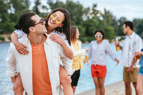 handsome young man piggybacking girlfriend while having fun on beach together with multicultural friends