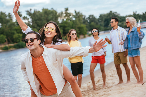 cheerful young man piggybacking happy girlfriend while walking on beach together with multicultural friends