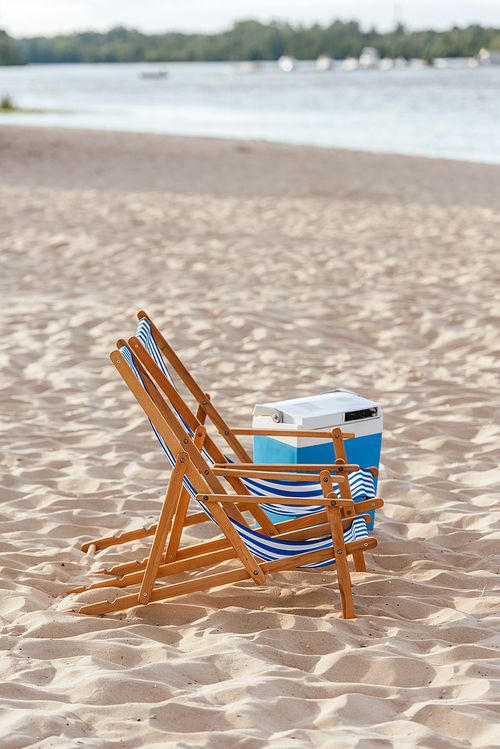 two chaise lounges and portably refrigerator on sand beach near river