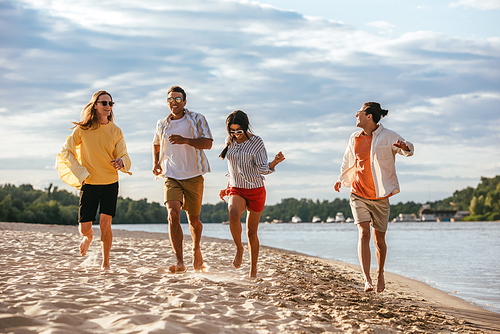 cheerful multicultural friends running on sand beach near river