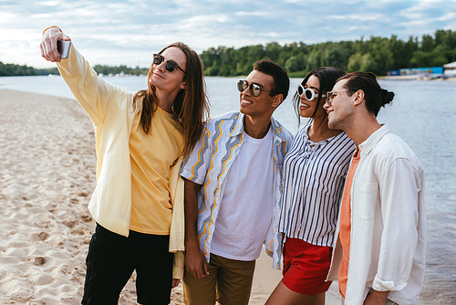 smiling young man taking selfie with cheerful multicultural friends on beach