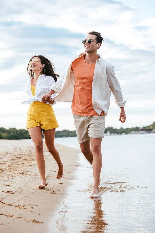 happy young couple holding hands while running on beach near river