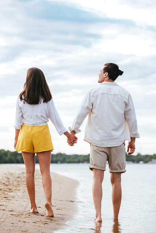 back view of young couple holding hands and looking at each other while walking on riverside
