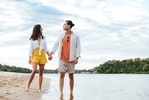 happy young couple holding hands and looking at each other while walking on riverside