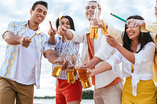 happy multicultural friends holding sparklers and bottles of beer while having fun on beach