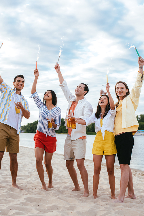 cheerful multicultural friends having fun on beach while holding sparklers in raised hands