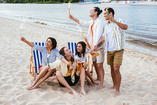 cheerful multicultural friends holding bottles of beer and sparklers while having fun on beach