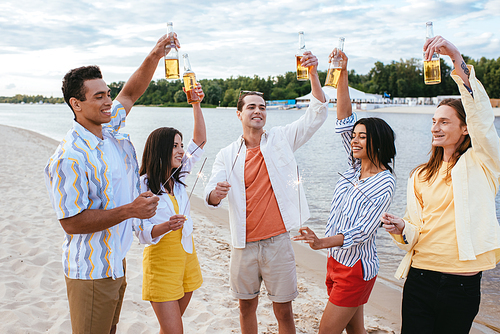 cheerful multicultural friends holding bottles of beer in raised hands and lighting sparklers on beach