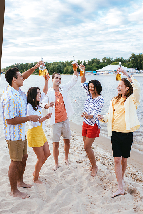 smiling multicultural friends holding bottles of beer and sparklers while having fun on beach