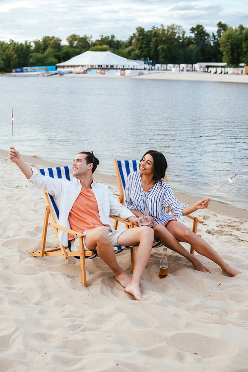 happy interracial couple sitting in chaise lounges and holding sparklers while resting on beach