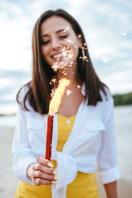 selective focus of happy young woman holding sparkler while standing on beach