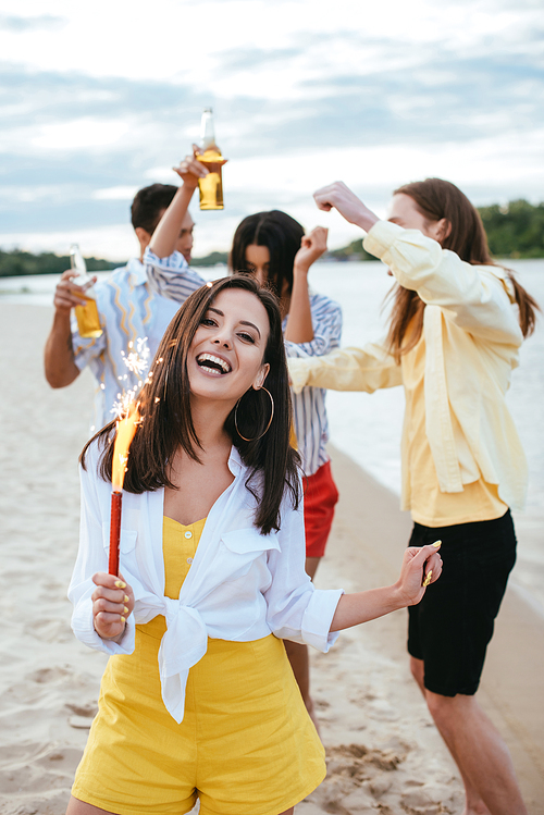 happy young woman holding sparkler and  while having fun on beach near multicultural friends