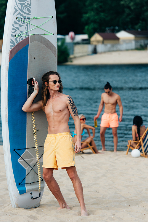 handsome young man looking away while standing near surfboard on beach