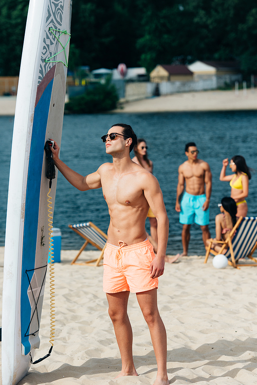 handsome young man in sunglases standing near surfing board on beach