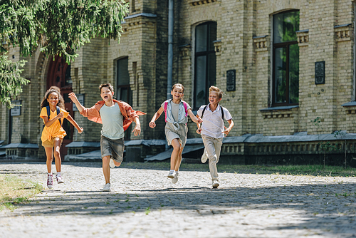 four adorable multicultural schoolchildren smiling while running in schoolyard