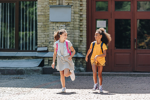 two cheerful multiethnic schoolgirls smiling while running in schoolyard