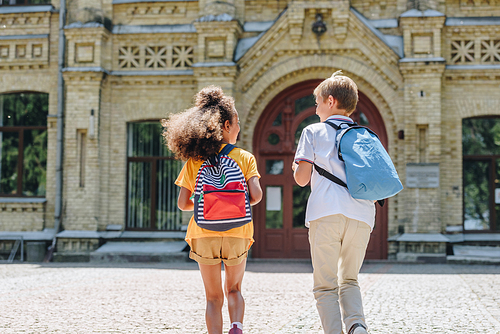 back view of two multicultural schoolkids with backpacks running in schoolyard