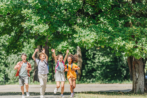 four happy multicultural schoolkids gesturing with raised hands while running in park