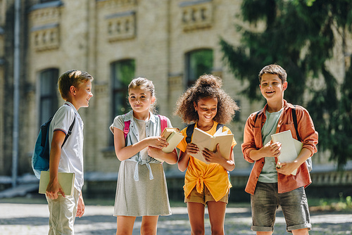 four adorable multicultural schoolchildren standing in schoolyard and holding books