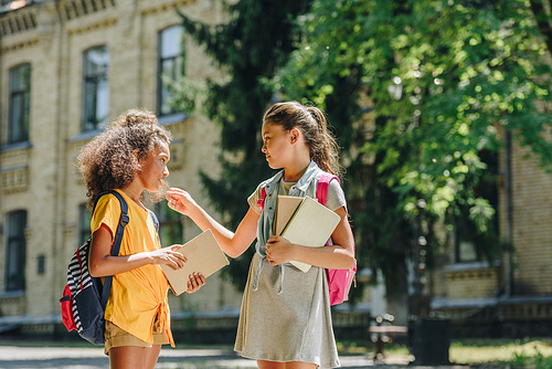 two multicultural schoolgirls holding books and speaking while standing in schoolyard