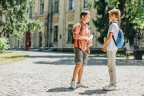 two cute schoolboys holding books and speaking while standing in schoolyard