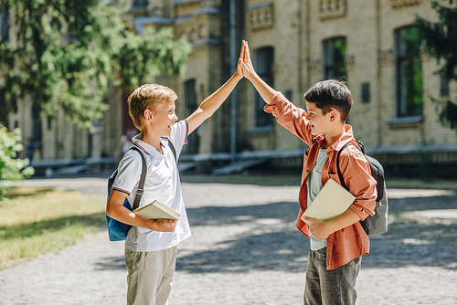 two cheerful schoolboys giving high five while standing in schoolyard