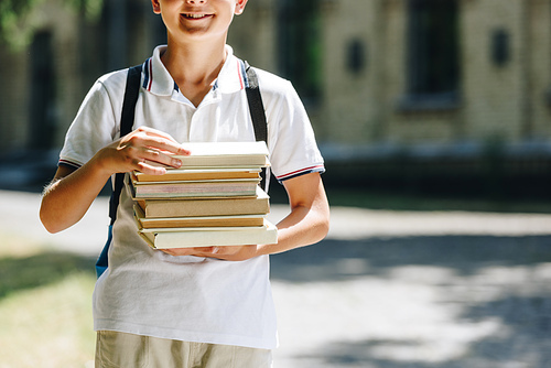 cropped view of smiling schoolboy with backpack holding books in schoolyard