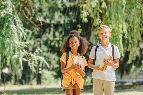 two cheerful multicultural schoolkids smiling at camera while holding books