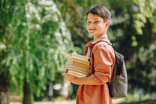 happy schoolboy smiling at camera while standing in park and holding books