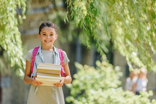 cheerful schoolgirl smiling at camera while standing in park and holding books