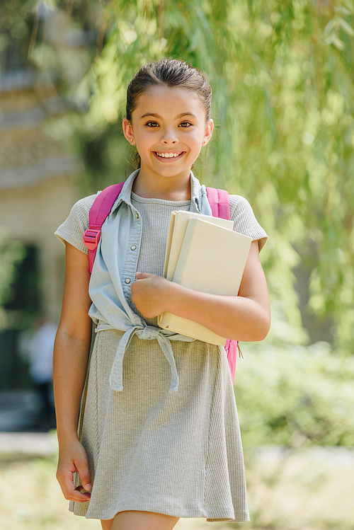 cute, smiling schoolgirl  while holding books in park