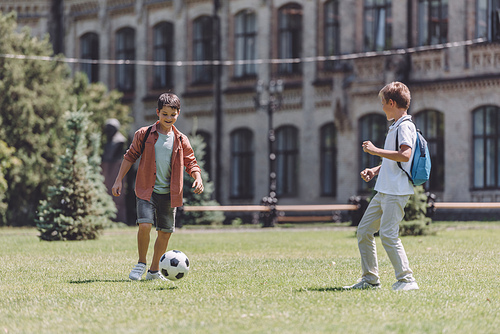 two cheerful schoolboys with backpacks playing football on lawn near school