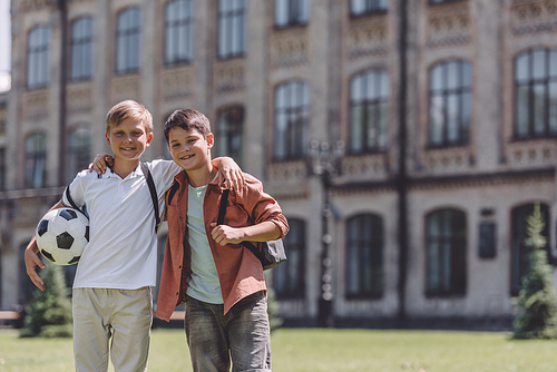 two cheerful schoolboys hugging while standing near school with soccer ball