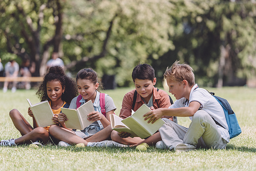 four adorable multicultural schoolkids sitting on lawn and reading books