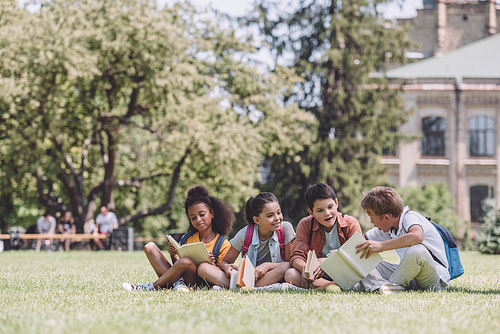 four cheerful multicultural schoolkids sitting on lawn and reading books