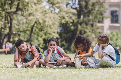 four bored multicultural schoolchildren with books sitting on lawn in park