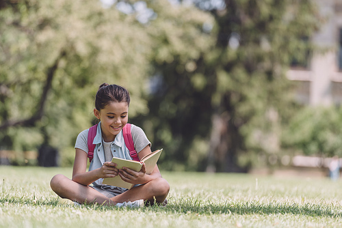 adorable, smiling schoolgirl sitting on lawn in park and reading book