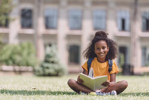cute african american schoolgirl smiling at camera while sitting on lawn with book