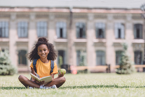 smiling african american schoolgirl  while sitting on lawn with book and holding apple