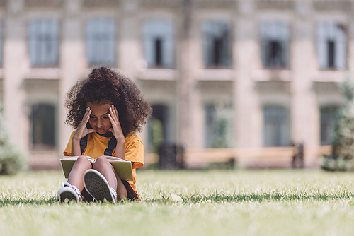 attentive african american schoolgirl reading book while sitting on lawn