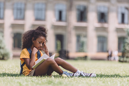 concentrated african american schoolgirl reading book while sitting on lawn