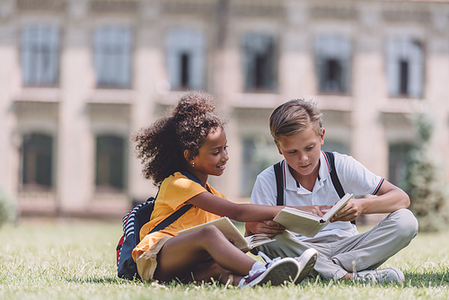 two adorable multicultural schoolkids sitting on lawn and reading books together