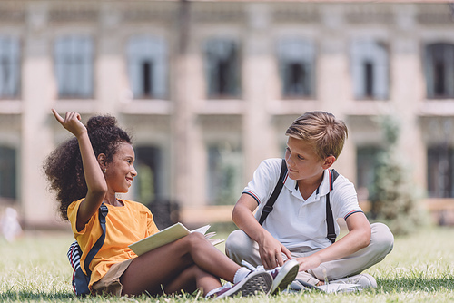 cheerful african american schoolgirl showing idea sign while sitting on lawn near multiethnic friend