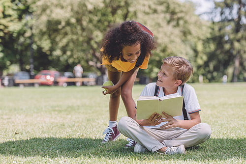 smiling african american schoolboy standing near multicultural friend sitting on lawn with book