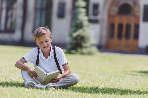 adorable schoolboy sitting on lawn near school and reading book