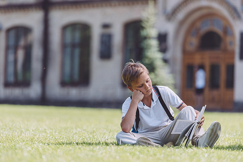 attentive schoolboy sitting on lawn near school and reading book