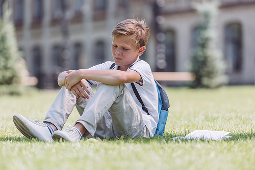 pensive schoolboy with backpack sitting on green lawn near school